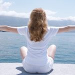 Woman sitting with open arms by the water, embracing nature and serenity.